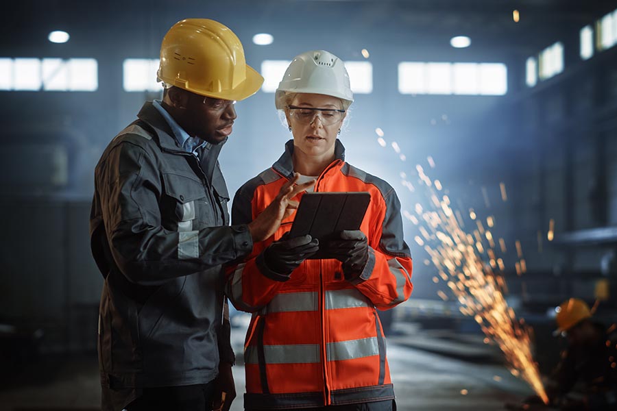 Specialized Business Insurance - Construction Workers Consulting Over a Tablet in a Warehouse With Metalwork Going on Behind Them